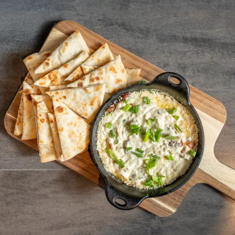 Creamy dip topped with green onions in a cast iron skillet, served with pita bread on a wooden board.