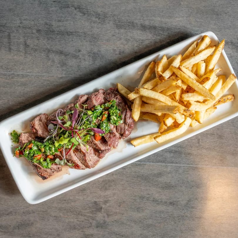 Sliced steak with green chimichurri sauce and microgreens, served with a side of golden french fries.