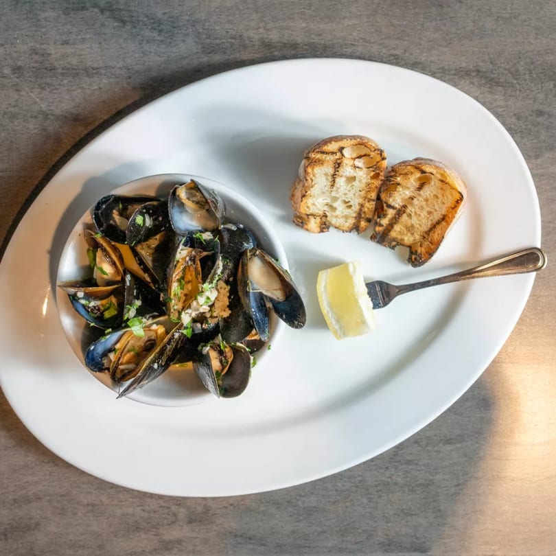 Steamed mussels in a white bowl with grilled bread and a lemon wedge on a white plate.
