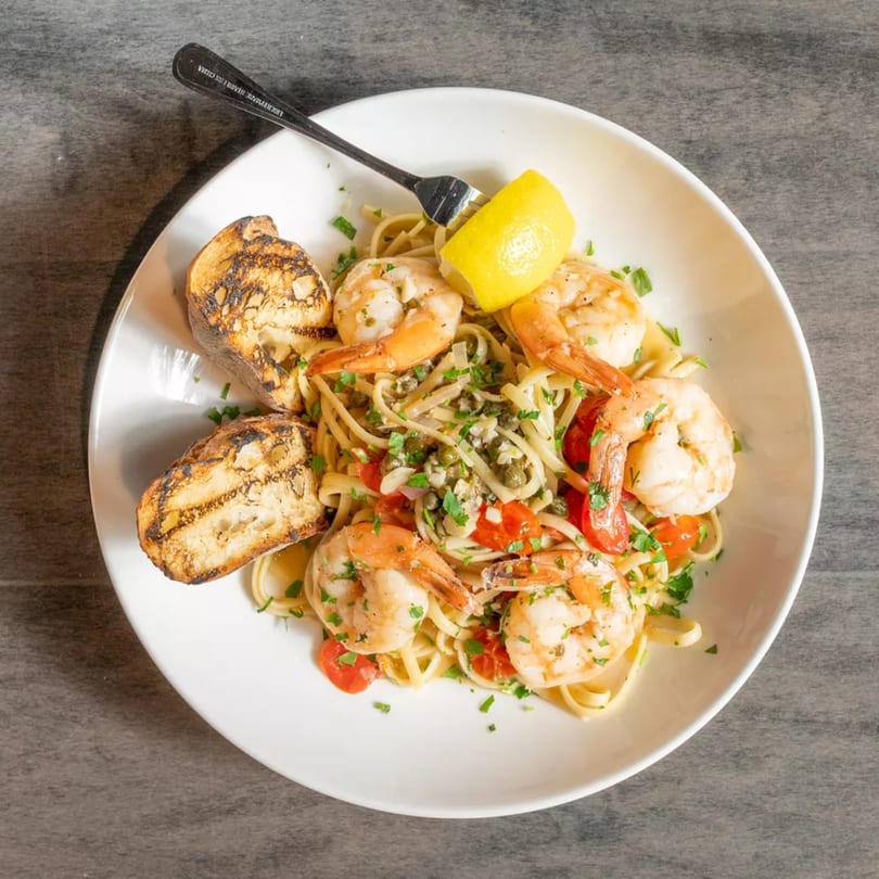 Shrimp scampi with linguine, cherry tomatoes, capers, parsley, lemon, and grilled bread on a white plate.