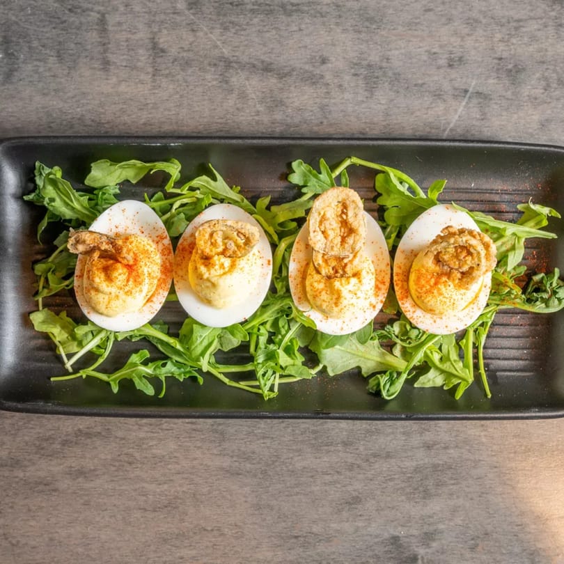 Four deviled eggs with fried oysters and paprika, served on a bed of arugula on a black plate.